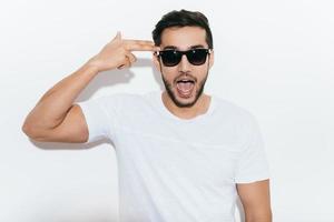 Bang Playful young Indian man in sunglasses gesturing handgun near head and making a face while standing against white background photo