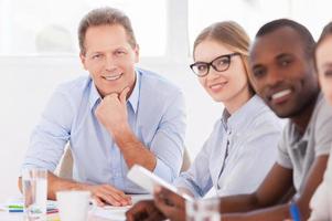 Strong and creative team leader. Confident mature man sitting at the table while his colleagues sitting in a row and looking at camera photo