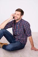 Friendly talk. Cheerful young man talking on the mobile phone and smiling at camera while sitting on hardwood floor photo