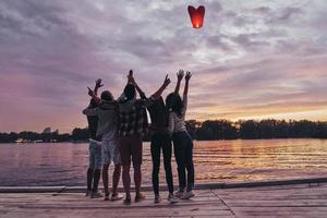 volando lejos... vista trasera completa de los jóvenes manteniendo los brazos levantados y mirando la linterna del cielo flotante mientras están de pie en el muelle foto