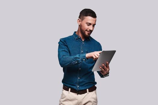 Surfing the net. Good looking young man in smart casual wear using digital tablet and smiling while standing against grey background photo