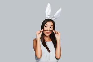 Enjoying carefree time. Beautiful young Asian woman in bunny ears looking away while standing against grey background photo