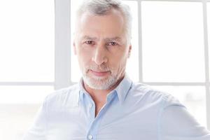 Confident and successful. Confident grey hair senior man in shirt looking ay camera while standing in front of the window photo