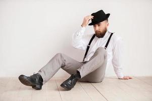 hombre guapo y confiado. un joven pensativo con un elegante sombrero informal en la mano y mirando hacia otro lado mientras se sienta en el suelo foto