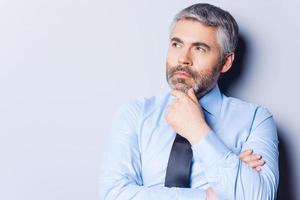 Lost in business thoughts. Thoughtful mature man in shirt and tie looking away and holding hand on chin while standing against grey background photo