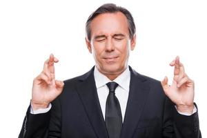 Waiting for special moment. Portrait of mature man in formalwear keeping fingers crossed and eyes closed while standing isolated on white background photo