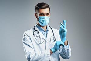 Serious young man in white lab coat and protective mask adjusting glove while standing against grey background photo