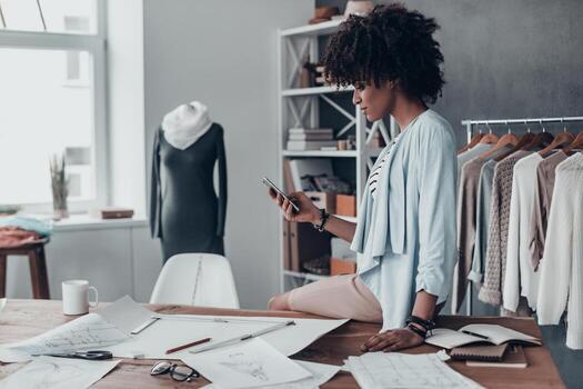 otro cliente una joven africana seria sosteniendo un teléfono inteligente y mirándolo mientras estaba sentada en el escritorio de su taller cerca de la ropa colgada en los estantes foto