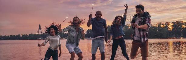 Feels like flying. Full length of young people in casual wear smiling and holding sparkers while jumping on the pier photo