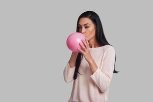 Time for fun Studio shot of attractive young woman in casual wear blowing up a pink balloon while standing against grey background photo