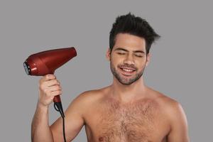 Hair is important part of his look. Handsome young man drying his hair and smiling while standing against grey background photo