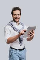 utilizando tecnologías modernas. un joven hermoso con ropa informal elegante que trabaja en una tableta digital y mira la cámara mientras se enfrenta a un fondo gris foto