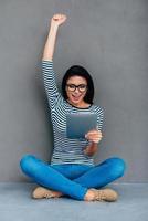 I did it Happy young woman holding digital tablet and keeping arm raised while sitting on the floor and against grey background photo
