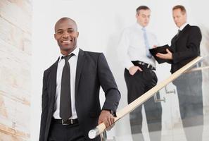 Confident businessman. Cheerful young black man in formalwear moving down by staircase and smiling while two people talking on background photo