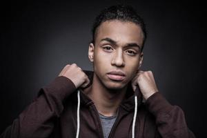 In my own style. Portrait of young African man looking at camera and adjusting his hood while standing against black background photo