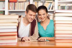 Studying together. Cheerful young man and woman sitting close to each other at the library desk and reading book photo