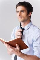 Just inspired. Thoughtful young man in formalwear holding note pad and touching his chin with pen while standing against grey background photo