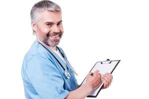 Writing RX prescription. Side view of mature doctor looking at camera and smiling while writing something at his clipboard and standing isolated on white photo