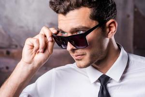 genial y guapo. joven confiado con camisa y corbata ajustando sus gafas de sol y mirando hacia otro lado mientras está de pie contra el fondo de metal foto