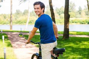 Enjoying great time in park. Rear view of happy young man riding bicycle in park and looking over shoulder photo