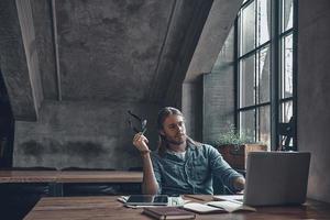 Putting ideas into something real. Thoughtful young man holding eyeglasses and working using computer while sitting at the office desk near the window photo