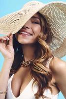 Portrait of beautiful young woman in elegant hat looking at camera and smiling while standing against blue background photo