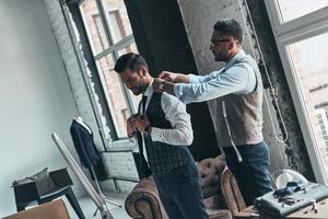 Thinking about every detail. Young fashionable designer helping his client to get dressed while standing in his workshop photo