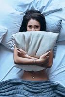 Just one more minute in bed Top view of attractive young woman covering half of her face with pillow and looking at camera while lying on the bed at home photo
