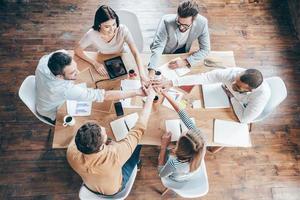 Starting new working day as a team. Top view of group of six young people holding hands together and smile while sitting at the office desk photo