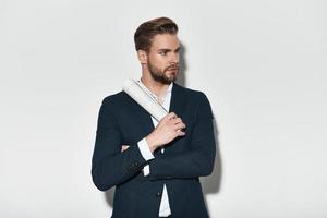 Confidence and charisma. Handsome young man in full suit keeping arms crossed and looking away while standing against grey background photo