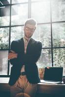 Young and successful. Confident young man in smart casual wear keeping arms crossed and looking at camera while standing in front of the big window in office or cafe photo