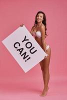 Full length of attractive young woman holding a poster and smiling while standing on tiptoe against pink background photo