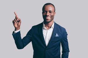 Pointing copy space. Handsome young African man in smart casual jacket pointing copy space and smiling while standing against grey background photo