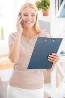 Sharing good news. Beautiful young woman leaning at the shelf in office and smiling while talking on the mobile phone and looking at clipboard photo