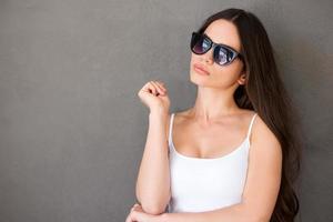 mirada de moda mujer joven confiada en anteojos mirando a la cámara mientras está de pie contra el fondo gris foto