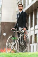Enjoying urban lifestyle. Low angle view of confident young businessman looking away and holding hands on his bicycle while standing outdoors photo