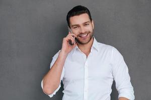 en el teléfono apuesto joven hablando por teléfono móvil y sonriendo mientras está de pie contra el fondo gris foto