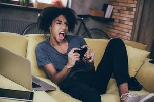 This game is so exciting Excited young African man playing video game and keeping his mouth open while sitting on the couch at home photo