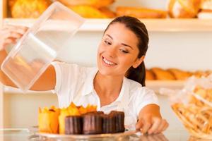 la panadería más fresca para nuestros clientes. hermosa joven en delantal llevando plato con galletas frescas y sonriendo mientras está de pie en la panadería foto