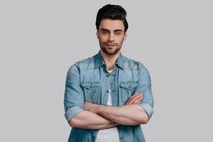 Portrait of confidence. Good looking young man in blue jeans shirt keeping arms crossed and looking at camera while standing against grey background photo