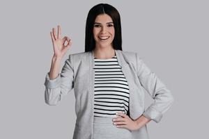 Everything is great  Beautiful young woman in smart casual wear gesturing and looking at camera with smile while standing against grey background photo