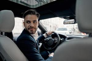 Driven by success. Handsome young man in full suit looking over shoulder and smiling while driving a car photo