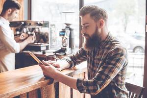 Working at coffee shop. Side view of young handsome man using his digital tablet while sitting at bar counter at cafe with barista at background photo