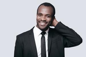 Handsome businessman. Portrait of confident young African man holding hand behind head and smiling while standing against grey background photo