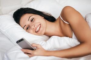 Texting to boyfriend. Attractive young woman smiling and holding smart phone while lying in the bed at home photo