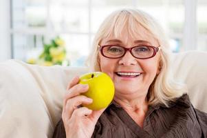 Enjoying healthy eating. Senior woman holding apple and smiling while sitting at the chair photo