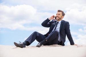 mantenerse conectado. feliz joven hombre de negocios hablando por teléfono móvil y sonriendo mientras se sienta en la arena y contra el cielo azul foto