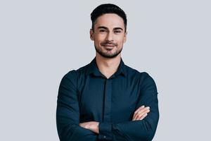 Young and successful. Handsome young man in shirt keeping arms crossed and smiling while standing against grey background photo