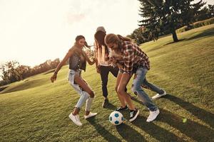 toda la longitud de jóvenes sonrientes con ropa informal disfrutando de un agradable día de verano mientras juegan al fútbol al aire libre foto