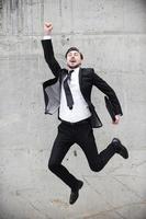 Great success. Happy young man in formalwear keeping arms raised and expressing positivity while jumping in front of the concrete wall photo